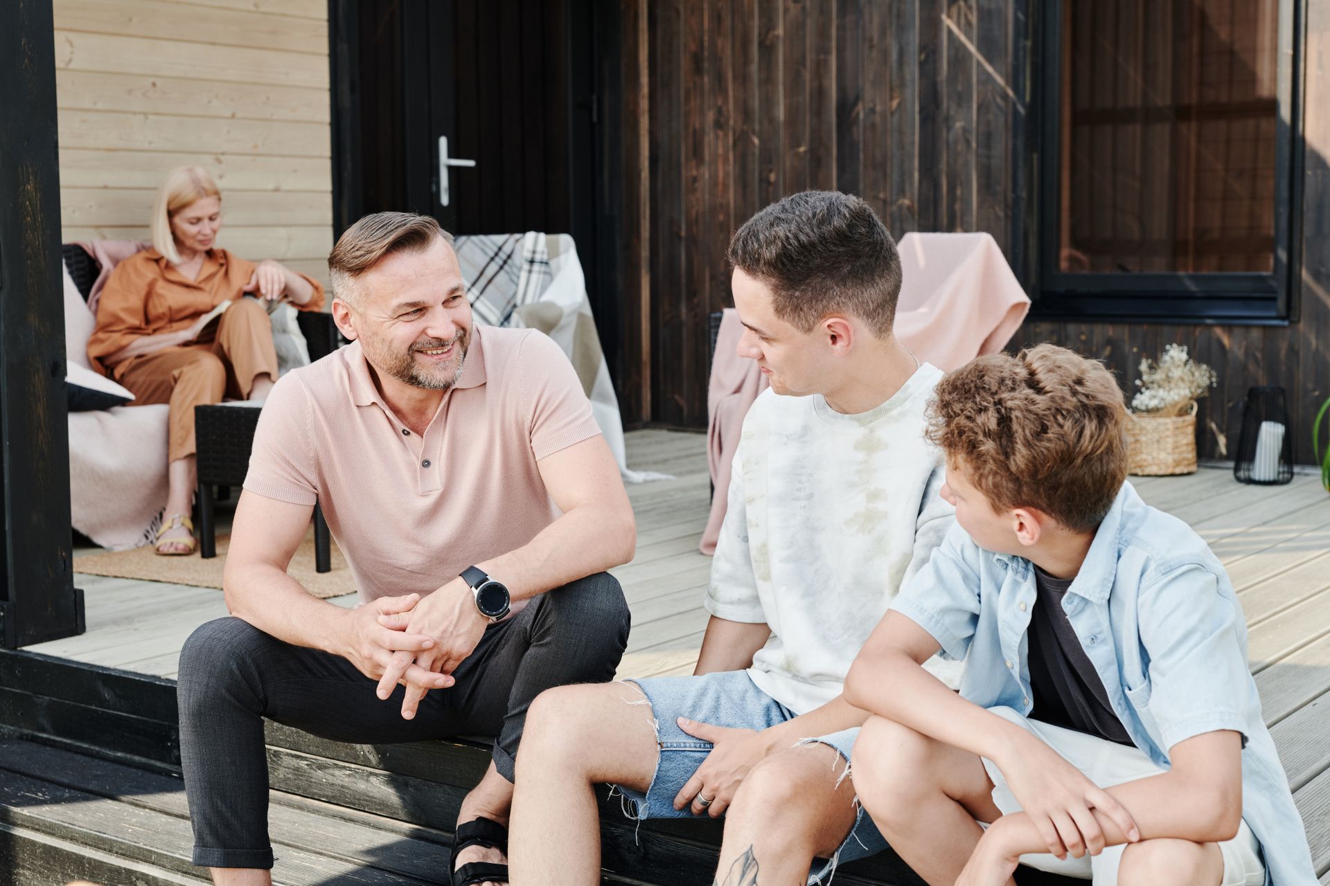 A parent laughs with their two children on a porch step, another parent reading quietly behind them.
