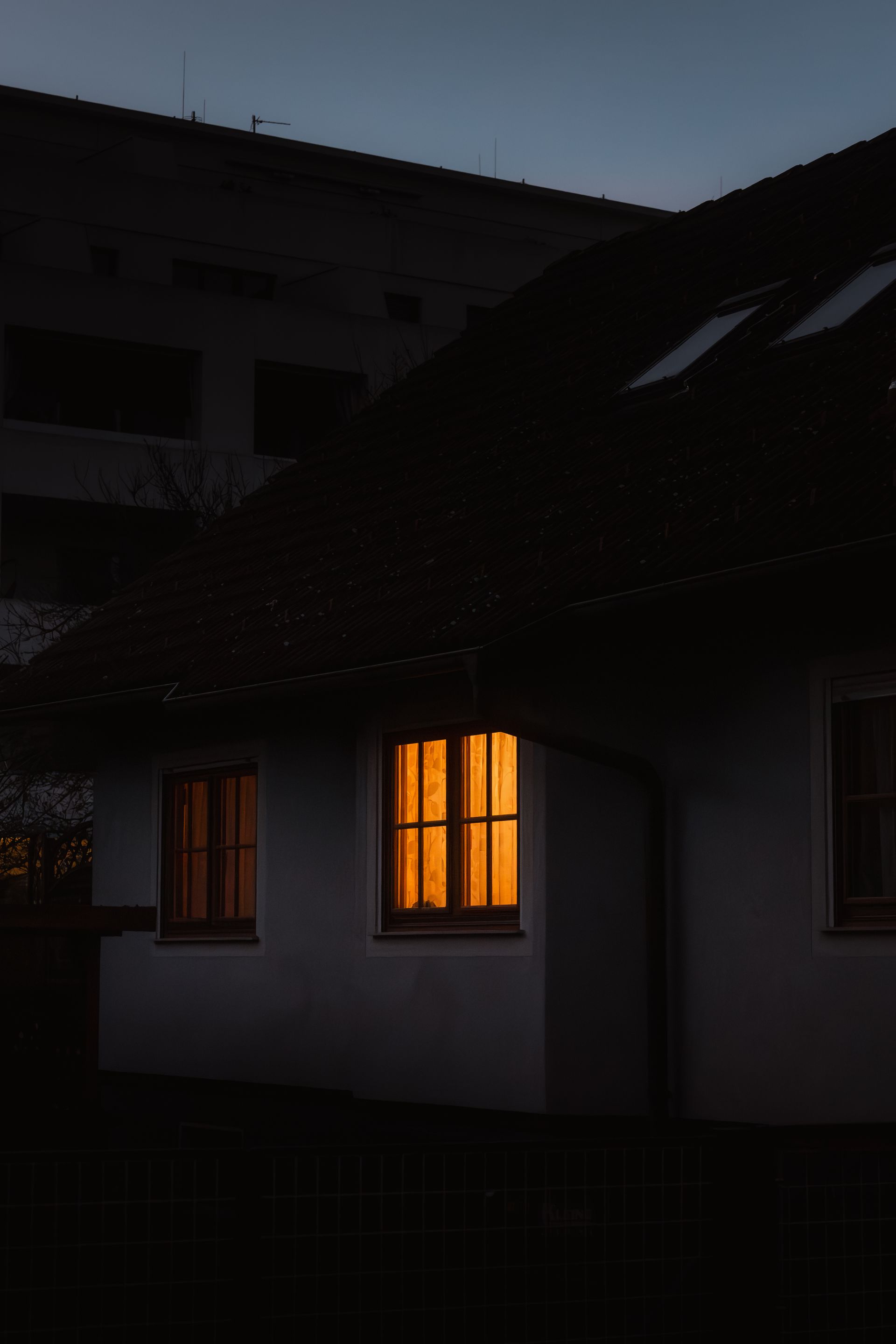 A single lit window glows warmly in an otherwise dark house at dusk.