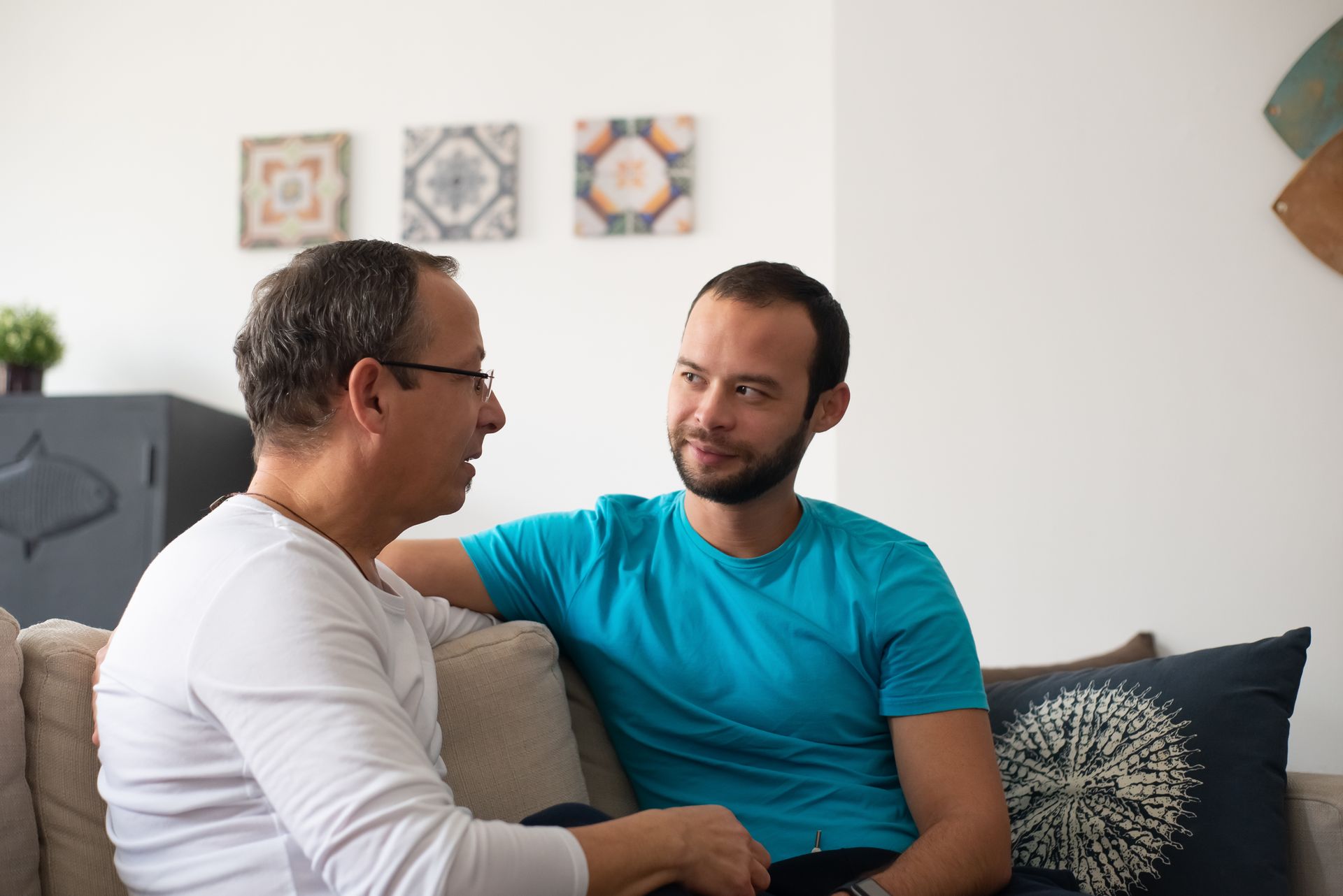 An older mentor and a younger person in an unhurried one-on-one conversation on a living-room sofa.