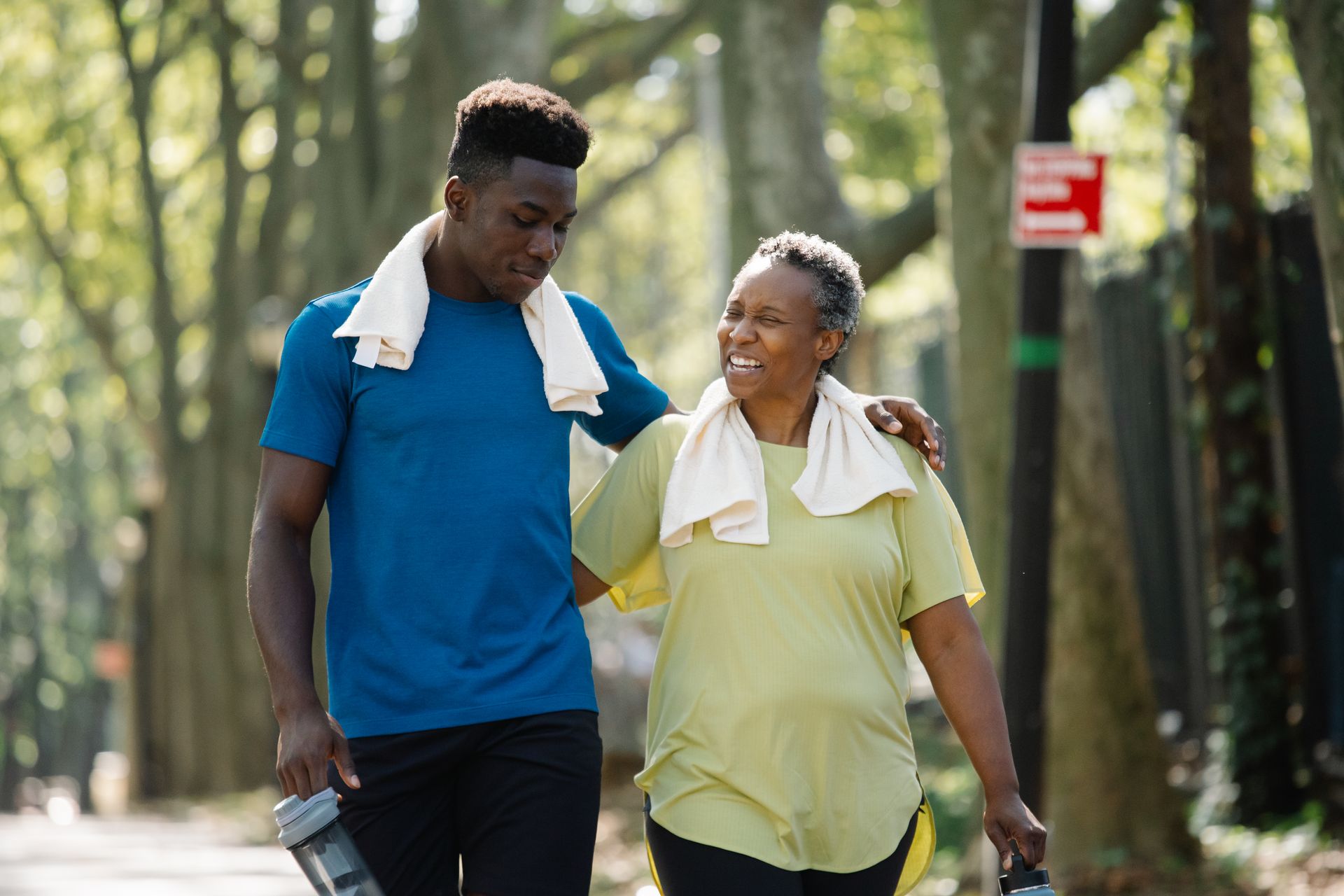 A parent and their adult child walk together along a sunlit, tree-lined path.